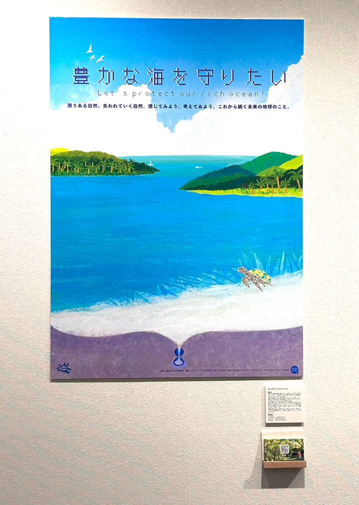 展示されている本山浩子の環境ポスター。美しいコバルトブルーの海と緑豊かな自然の島々、晴れた青い空に羽ばたく鳥、白い砂浜とウミガメのほのぼのした優しいイラストです。